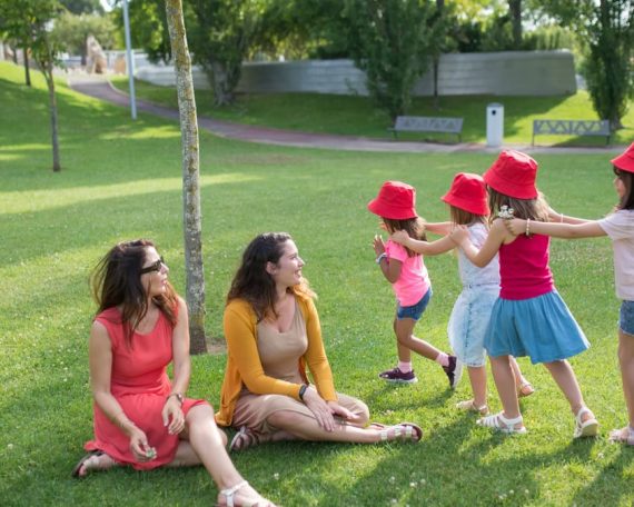 Happy children playing in a sunny park with two mothers sitting nearby, enjoying the outdoors.